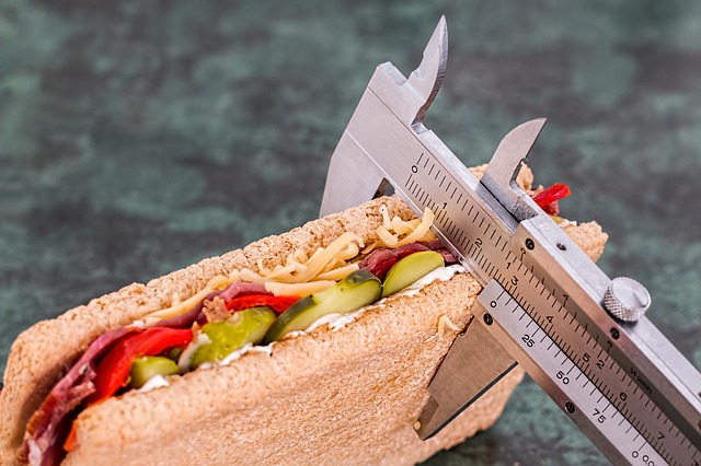 A metal sliding scale measures the thickness of the sandwich. The sandwich is sliced and filled with cucumbers, red peppers, salami and grated cheese.